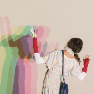 A woman stands in front of a white wall. A light shines on her from behind, casting a multi-coloured shadow on the wall.