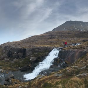 A panorama from the base of Beinn Eighe, looking north. In the right of the frame a small waterfall flows left. Chris is stood at the top of the waterfall in a bright red jacket.