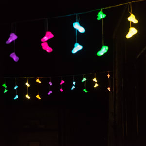 Pairs of brightly coloured lights in the shape of shoes dangle from a line suspended between two rows of houses.