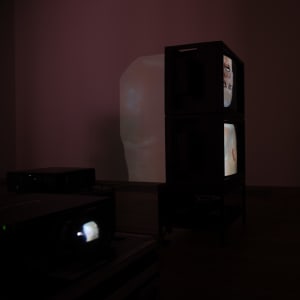 Two vintage television monitors are stacked vertically in a dark room, each showing a bald man’s head. The same head is projected on the wall behind them.