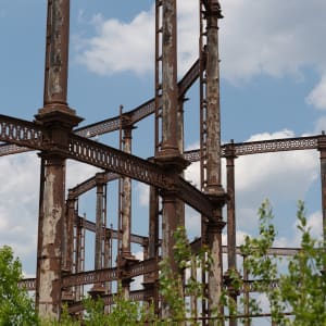 A portrait photo of the side of a gas works, with another visible in the distance. The metal is mostly rusted with patches of peeling paint visible.