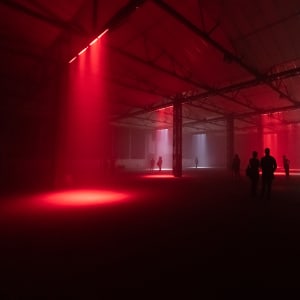 A cavernous dark warehouse. Several lights can be seen pointed at the floor like spotlights - some red and some white. Several figures can be seen silhouetted in the dark.