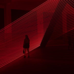 Red lasers in a dark room form even horizontal lines going from bottom left to upper right. A figure is silhouetted in front of them.