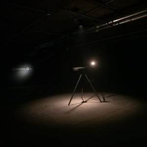 In a pitch black room a tripod can be seen in the centre illuminated by a single point of light on top of the tripod facing towards the camera.