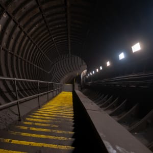A staircase descends a dimly lit tunnel. There are regular dim lights on one side and the stair treads are painted yellow.