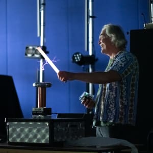 Man in a dark room holding a 2′ fluorescent tube in front of a small tesla coil. The tube is lit up and there are purple sparks from the coil to the tube.
