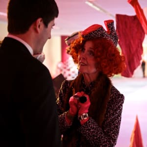 A woman wearing a shoe-hat talks to a man.