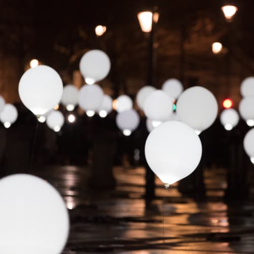 Many balloons float above a dark street - the balloons are lit like light bulbs.