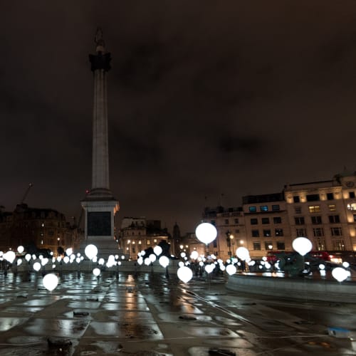 Many balloons float above the ground of Trafalgar Square - the balloons are lit like light bulbs.