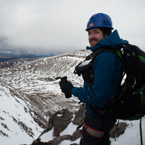 Ed drinking from a vacuum flask, with snowy mountainside in the distance. He’s wearing full climbing gear - helmet, harness, rucksack.
