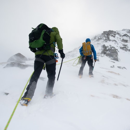 Ed and a guide walk up a snowy mountain on an overcast day. The camera angle is near to the ground looking up at their backs. They’re both connected by a climbing rope.