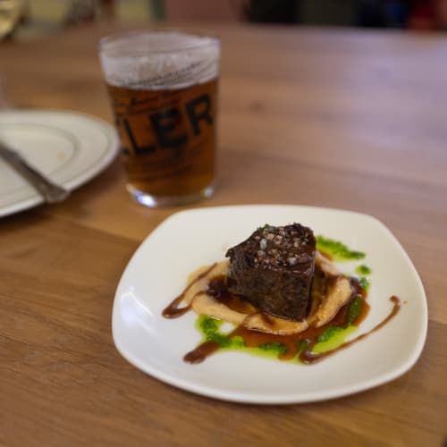 A small piece of beef sits on a plate green vinaigrette and a glass of beer in the background.