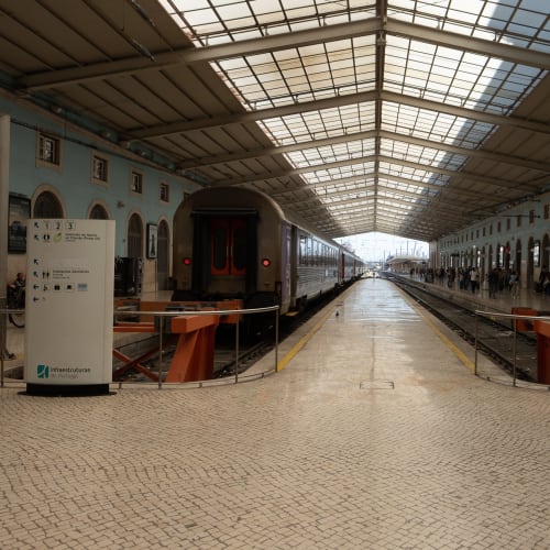 Looking directly down the central platform in a train station. On the left a train is waiting. The station is small and with a low roof.