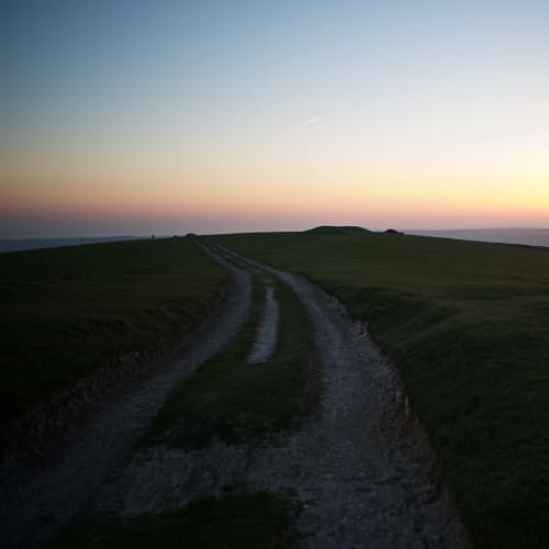 A chalky path recedes in to the distance on a hillside. The sun is just setting on the horizon.