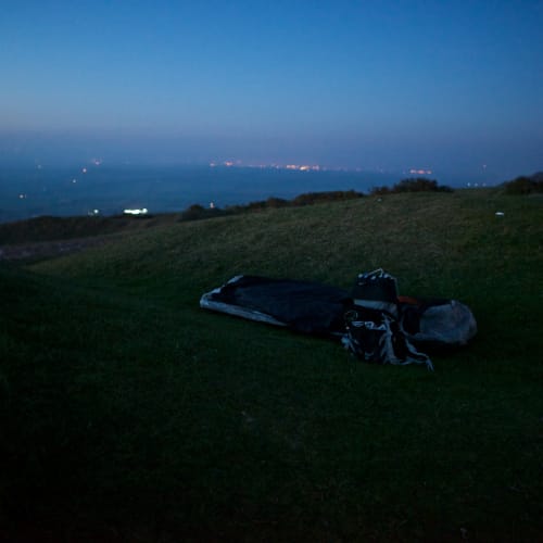A bivvy bag and rucksack sit in a hollow depression in the grass in the last of the evening light. The sky is dark blue and calm.