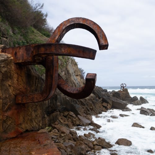 A photo of two parts of a large iron sculpture by artist Eduardo Chillida. The sculptures sit on the coastline with the sea to the right. One piece dominates in the foreground, with another just visible in the background.