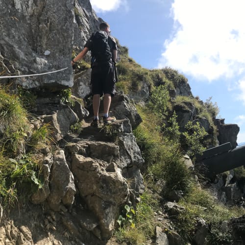 Oj climbing a steep path on the left, with a panorama of the alps to the right of him. In the center of the frame is a large pipe emerging from the hillside.