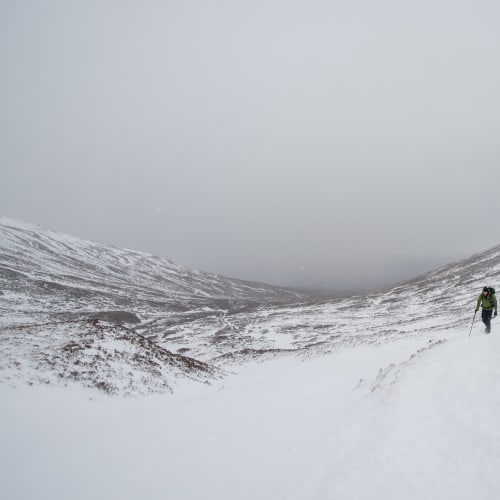looking down a snowy hill with low grey clouds. Ed is off to one side walking up towards the camera.