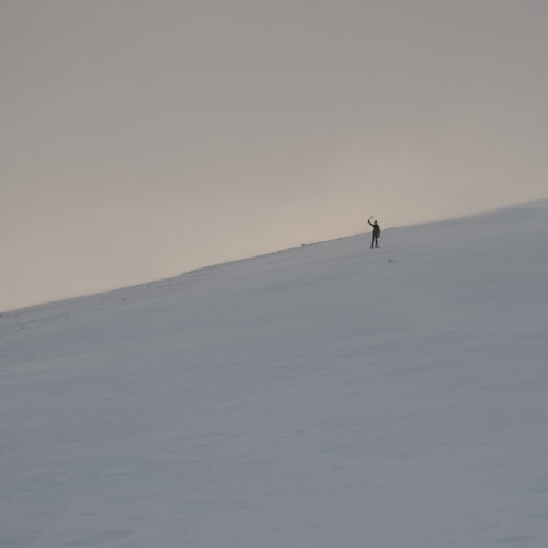 A wide shot at dusk looking up a snowy hill. A figure (Ed) in the distance can be seen waving an ice axe.