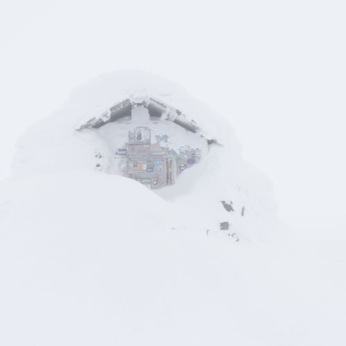 The emergency hut at the top of Ben Nevis. It’s covered in lots of snow, so only a patch of the top of the doorway is visible. There’s low visibility so it looks somewhat ghostly.