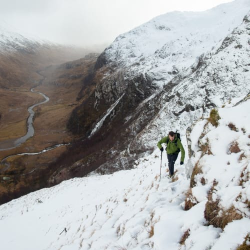 Ed climbing a snowy and grassy mountainside. In the far distance the brown base of the valley can be seen with a river running through the middle.