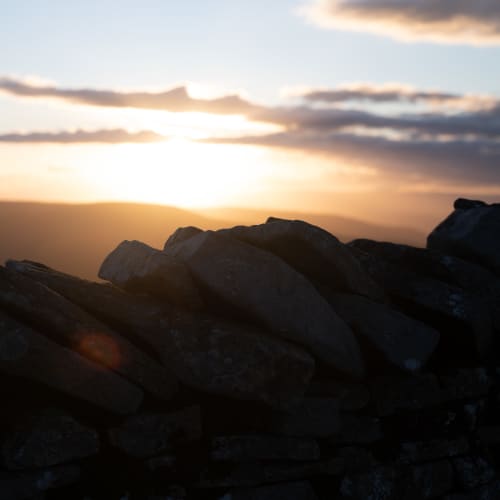A close photo of the top row of stones of a stone all. The stones all lean to one side. In the background the sun is just setting.