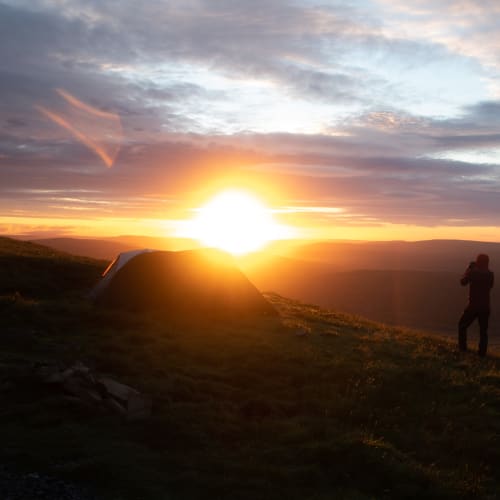 Sunrise over Whernside. Chris stands in the foreground in silhouette, taking a photo of the sun.