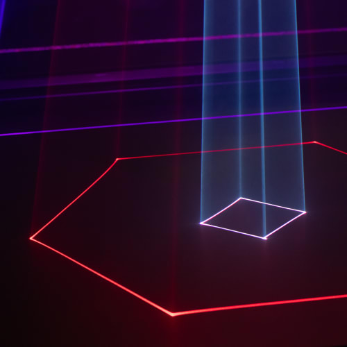 A laser beam projects shapes from above on to the ground. It has traced a large red hexagon, with a small pink square inside.