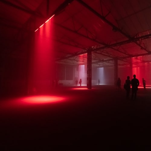 A cavernous dark warehouse. Several lights can be seen pointed at the floor like spotlights - some red and some white. Several figures can be seen silhouetted in the dark.