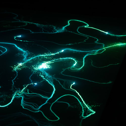 A photo from a moving light installation. A bright point light is projected on the floor, and wanders randomly around. The floor is covered in a material that glows for a period when exposed to light. The end effect is of random lines of light green and blue drawn all over the ground.
