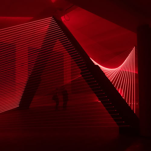 Red lasers in a dark room form lines that sweep up to a column then down again like threads.
