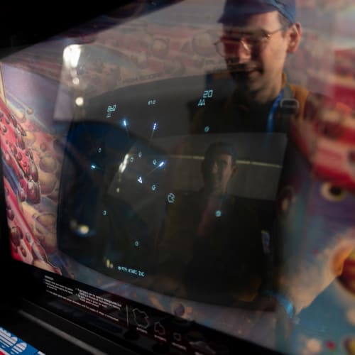 Close up photo of the screen of a vintage Asteroids arcade game with dark black screen and white ship and asteroids. There’s strong reflections on the cover glass and the player’s face is reflected twice.