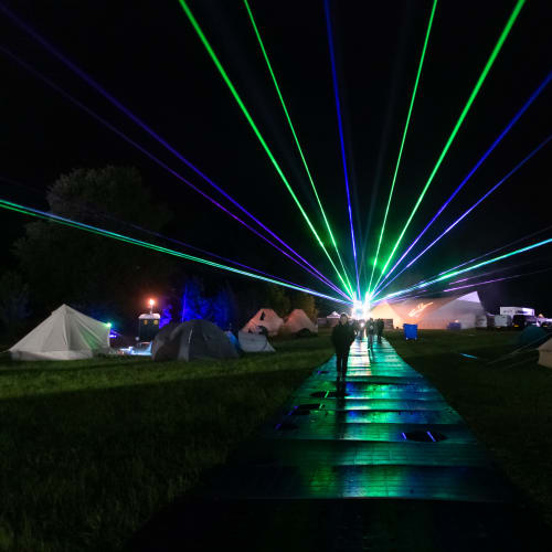 Looking straight down a path at night with white tents on either side. In the distance are lasers starting at the end of the path and going out in all directions in the sky.