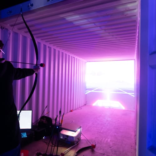 Person aiming a bow and arrow in to a shipping container with large screen at the far end.