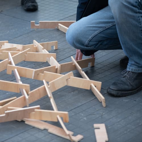 A person kneels next to a bunch of wooden slats connected together in a lattice