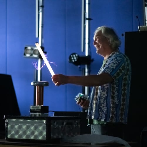 Man in a dark room holding a 2′ fluorescent tube in front of a small tesla coil. The tube is lit up and there are purple sparks from the coil to the tube.