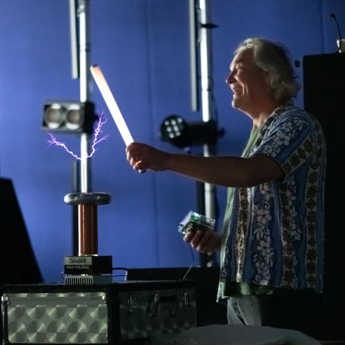 Man in a dark room holding a 2′ fluorescent tube in front of a small tesla coil. The tube is lit up and there are purple sparks from the coil to the tube.
