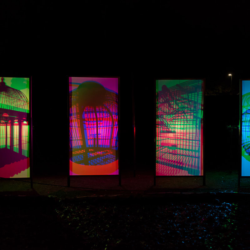 Four vertical panels are side by side in the dark. They’re light from behind with bright neon colours, projected with shadows of birdcages.