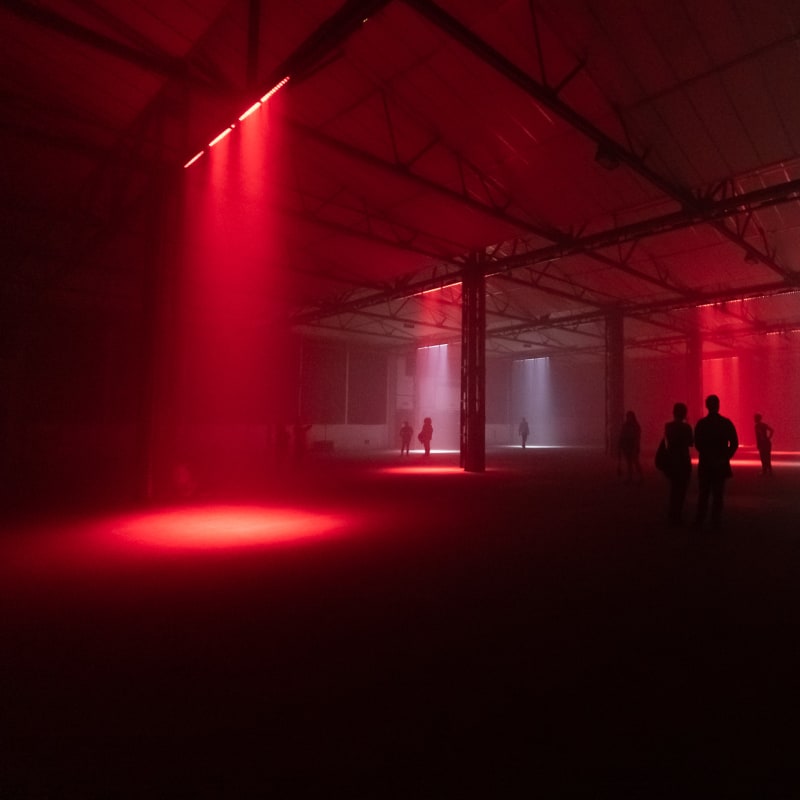 A cavernous dark warehouse. Several lights can be seen pointed at the floor like spotlights - some red and some white. Several figures can be seen silhouetted in the dark.