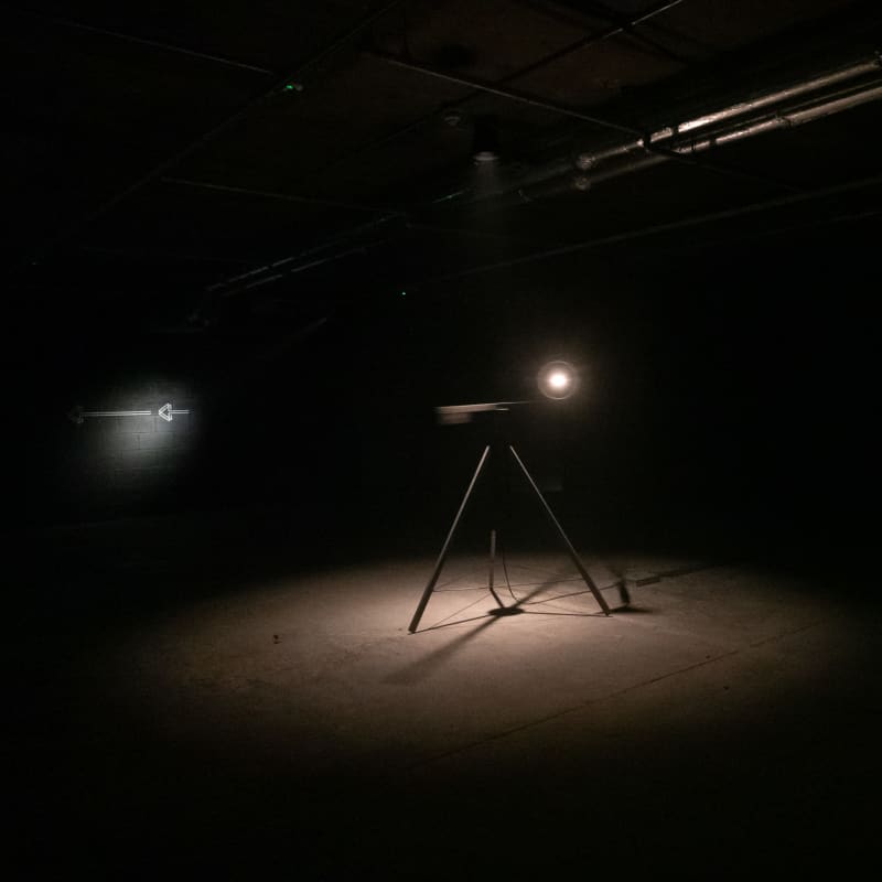 In a pitch black room a tripod can be seen in the centre illuminated by a single point of light on top of the tripod facing towards the camera.