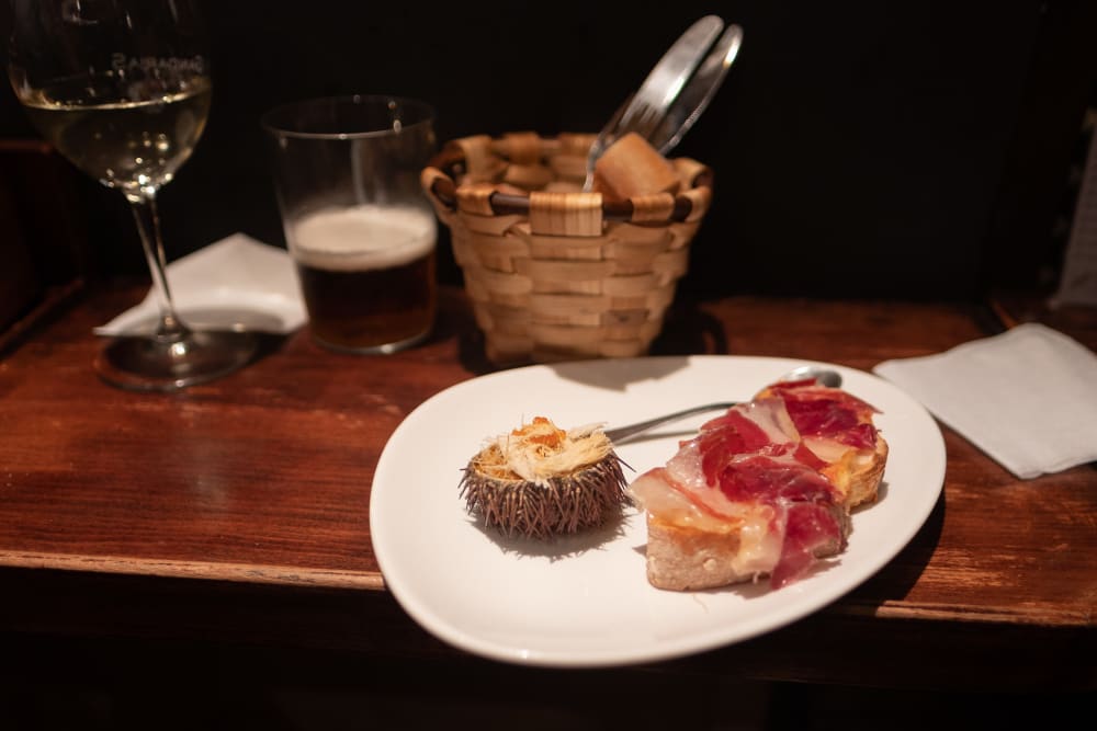 A plate of pintxos on a wooden bench next to a basket of bread, a glass of beer, and a glass of white wine. One pinxto is a slice of baguette with ham. The other is half of a sea urchin.