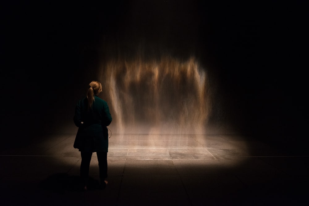 A fine mist of water drops in a dark room, lit from the side. A woman stands in front of the artwork.