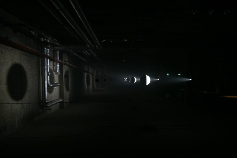 A pitch black room. A series of small white spheres are in the centre in a line to the distance. They are faintly light from the right casting shadows on the left wall. The White spheres look like moons.