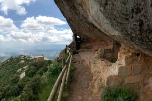 A panorama half looking over the Montserrat countryside and half looking at a path along the side of a mountain that is covered by steep rock.