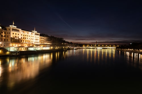 A panorama taken from a bridge over the Rhine at nighttime. There are buildings in the distance lit up and lights reflected in the water.