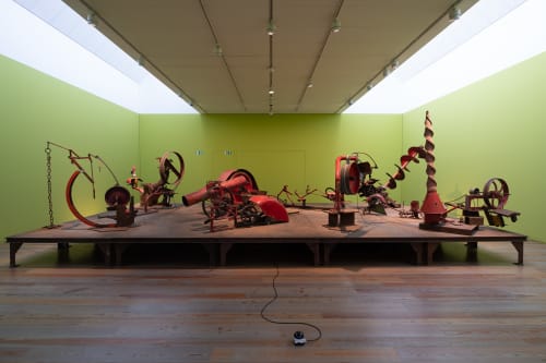 Wide photo of pale green room with several red mechanical sculptures in the middle.