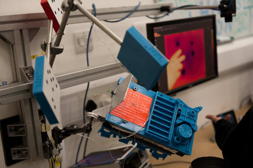 A photo of a science experiment. There are three blue stands held in the air. There’s a screen in the background with a hand visible on it.