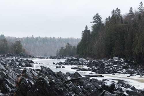 A long view across an icy river. There’s dark rocky outcrops coming out of the river. The river is partially frozen with creamy patches of ice at the sides.