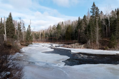 A photo down the banks of the Baptism river. The river is 70% covered in snow and ice, with a wire bridge in the background crossing the river.