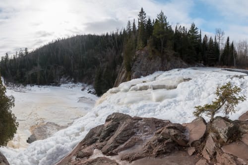 A panorama taken from the top of High Falls. The falls are partially frozen over with white and frothy ice.